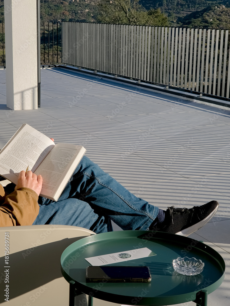 Obraz premium Man relaxing on a terrace esplanade reading a book during sunny winter afternoon, enjoying leisure time with stunning mountain landscape view from modern balcony