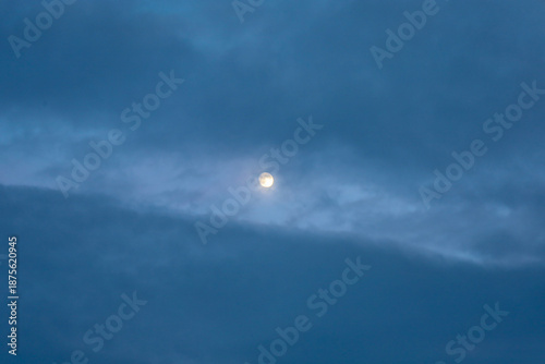 Konstfotografi A bright incomplete moon breaks through dense layers of blue evening clouds