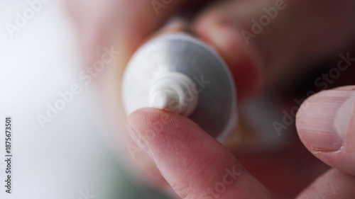 Close-up of hands squeezing a small amount of white cream from a tube onto a fingertip, illustrating skincare application, moisturizing routine, or topical treatment against a soft backdrop