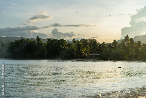 View of the tranquil ocean meeting the lush green shoreline under a sky streaked with golden sunlight and soft, puffy clouds, Vanimo, Sandaun Province, Papua New Guinea.