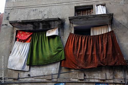View of weathered building facade with vibrantly colored curtains hanging from windows, casting shadows on the aged walls, Lahore, Punjab, Pakistan.