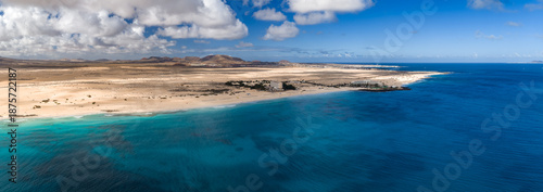 Fotografi Aerial view shows Playa del Bajo de la Burra in Fuerteventura, pale gold dunes, crescent shore, turquoise shallows to deep Atlantic, volcanic hills, resorts, palms, midday light