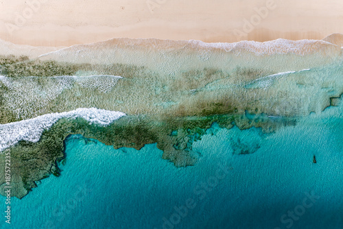 Aerial top down view shows pale sand, turquoise water, and a wide shallow reef near Playa del Bajo de la Burra or Morro Jable, with shorebreak lines and seagrass.
