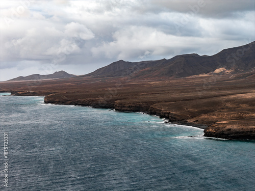 Tableau sur toile Dark lava cliffs and rust colored slopes meet the turquoise Atlantic in Fuerteventura, Canary Islands