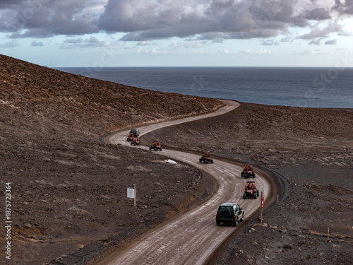 Slika na platnu A convoy of off road buggies and a black SUV winds a dusty coastal track in Fuerteventura, Canary Islands, with Atlantic Ocean under clouds and late day light