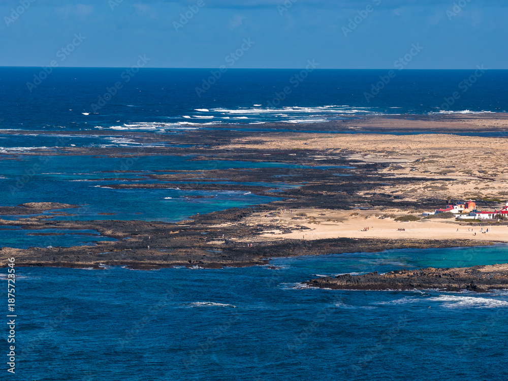 Obraz premium Aerial view shows Fuerteventura north coast, Playa del Bajo de la Burra, turquoise lagoons, volcanic reefs, whitewashed buildings, choppy Atlantic swells at midday.