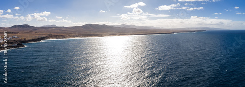 Aerial view shows Fuerteventura northwest coast at midday, a whitewashed village on a rocky headland, long beaches, cliffy points, and caldera silhouettes.