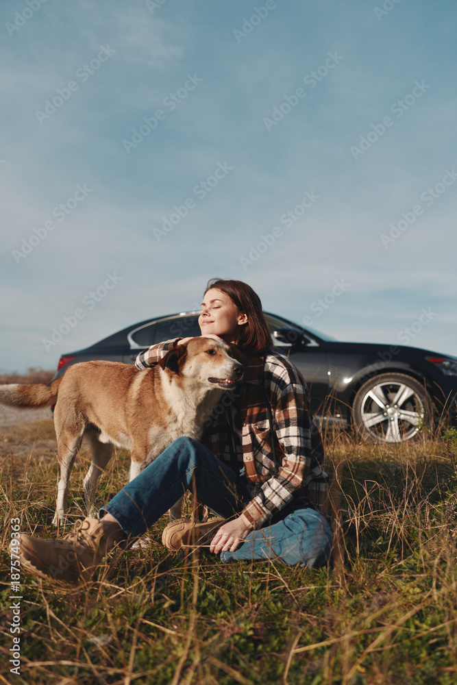 Fototapeta premium Woman sitting in a field with her dog, relaxing outdoors beside a car under a clear sky. Casual plaid shirt and jeans, a peaceful moment of companionship and nature on a roadside escape