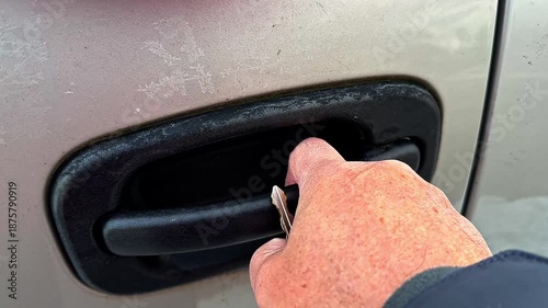 A hand is inserting a key into the door handle of a car in a parking lot. The weather is clear and sunny winter morning.