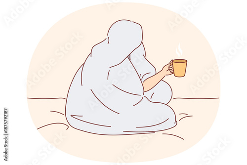 Man drinks coffee sitting on bed and wrapped in blanket to warm up after walk along winter street
