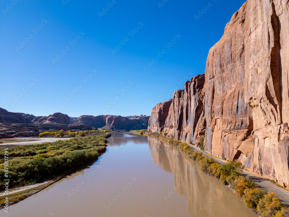 Fototapeta premium Aerial drone view of Wall Street's red sandstone cliffs in Moab at sunrise. The Colorado River winds alongside Potash Road as golden light hits climbers on the vertical walls and dark desert shadows