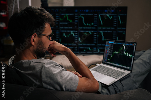 Canvas Print Thoughtful trader male in glasses focused on monitoring complex financial market data and cryptocurrency trends on laptop and large multi-screen setup, actively managing investments