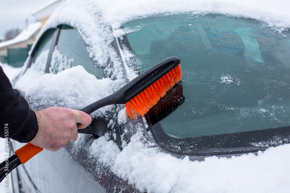 Fototapeta premium Hand holding a snow brush, clearing snow from a car window, showcasing winter weather conditions and the importance of vehicle maintenance during cold seasons