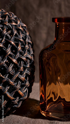 Dark still life with woven decorative object and amber glass bottle creating moody atmosphere luxury interior styling and artisanal design concept
