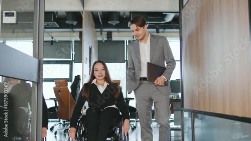 Two coworkers interact in a modern office. One person uses a wheelchair while discussing work matters with another in business attire.
