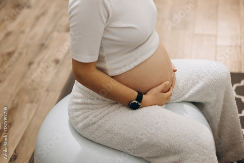 Pregnant Woman Sitting On A Fitness Ball At Home, Gently Holding Her Belly. Concept Of Maternity, Pregnancy Wellness, Prenatal Care, Self-Care, And Healthy Lifestyle. Intimate And Serene Indoor Scene 