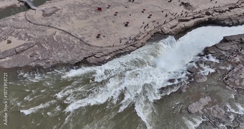Aerial video of Hukou Waterfall, mighty Yellow River thunders through a dramatic gorge with tourists along viewing paths on the Shanxi-Shaanxi border, China.