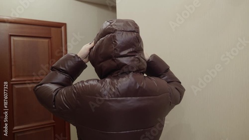 Adult woman standing indoors putting hood on brown winter jacket before leaving home. Cold season clothing and daily routine concept.