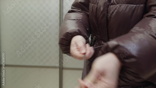 Adult woman tightening waist belt on brown winter jacket indoors. Cold season clothing, fit adjustment and daily routine concept.