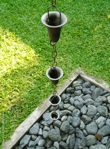 Zen-style functional garden decor featuring a decorative metal rain chain drainage system. Elegant outdoor landscaping detail with smooth river rocks and vibrant green grass background. Perfect for ho