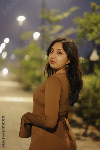 Stylish young woman wearing a brown long maxi dress posing confidently on an illuminated urban walkway at night