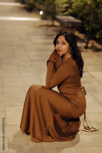 Stylish young woman wearing a brown long maxi dress posing confidently on an illuminated urban walkway at night