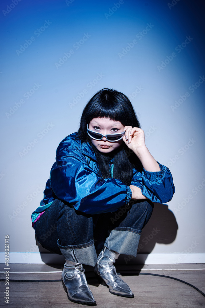 © Seventyfour - Asian young adult woman crouching near wall, holding sunglasses and looking directly at camera, wearing metallic boots, embodying second hand fashion style in urban setting © Seventyfour - Asian young adult woman crouching near wall, holding sunglasses and looking directly at camera, wearing metallic boots, embodying second hand fashion style in urban setting
