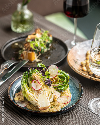 Принти на полотні A professional food photography shot of a grilled romaine lettuce heart served on a creamy base, garnished with sliced radishes and microgreens, with a glass of red wine in the blurred background