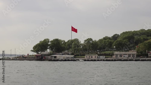 A Turkish flag waves atop a pole on a tree-lined shore beside calm waters.