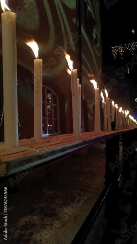A long row of white lit candles flickers on a wooden outdoors at night.Soft warm light illuminates the dark winter atmosphere with golden flames dancing against a blurred architectural background