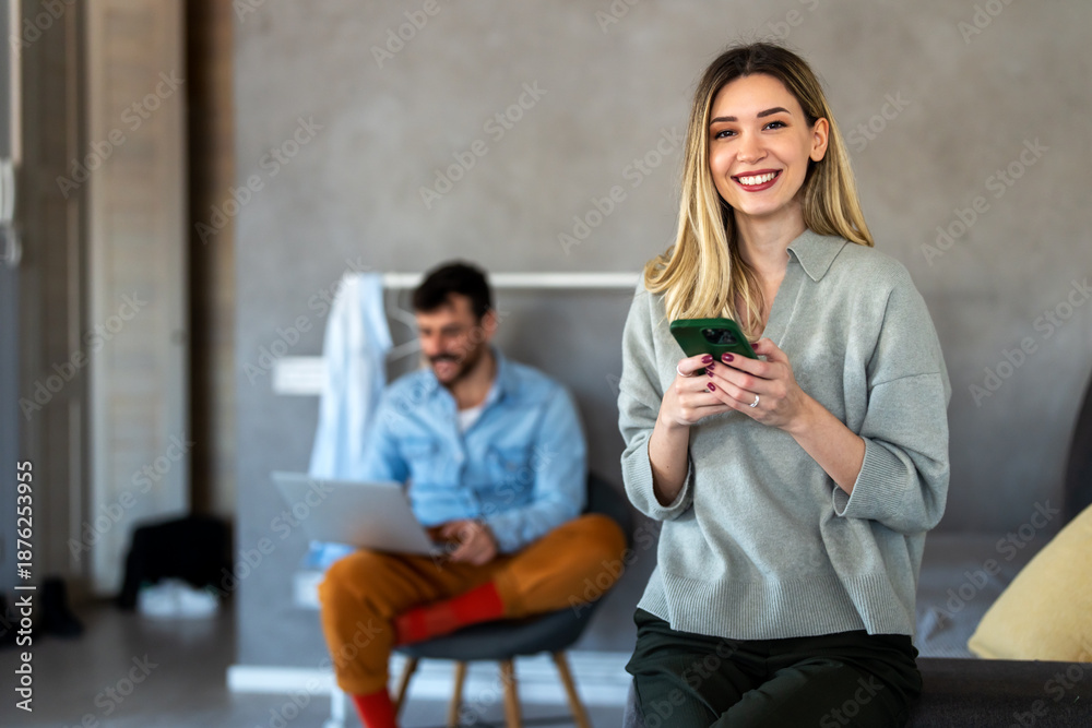 Fototapeta premium Smiling caucasian woman using her smartphone while her partner works in the background, home office