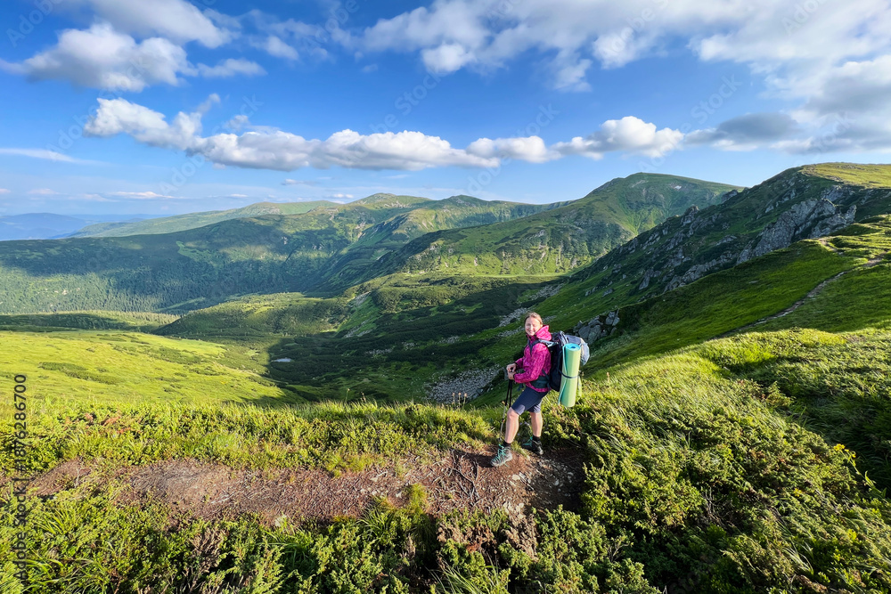Fototapeta premium Woman hiker stands on mountain trail, taking in breathtaking view of rolling green hills and valleys. Backpack and sleeping pad visible as she grips trekking poles under partly cloudy sky.