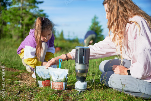 Fényképezés Tourist woman and young girl preparing meal from packaged pouch using portable camping stove on grassy hillside