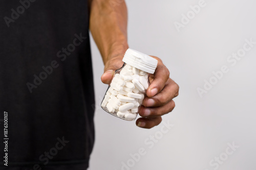 Man in black shirt extends clear bottle filled with small white oval supplement tablets toward the viewer.