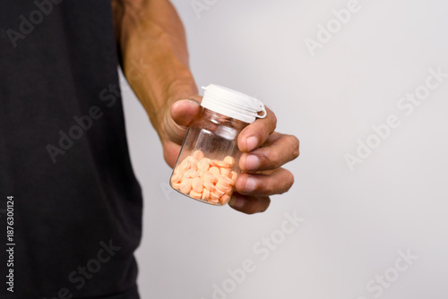 Close up shot shows a person's hand holding out a clear plastic prescription pill bottle filled with small orange tablets.
