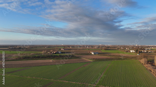 Po Valley agricultural landscape showing vast patchwork fields of winter crops and fertile soil. With scattered rural houses and distant towns under a partly cloudy sky