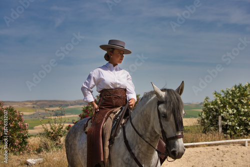 A young, beautiful Spanish woman dressed in riding clothes and wearing a traditional Spanish hat, riding a horse in the countryside on a sunny day.