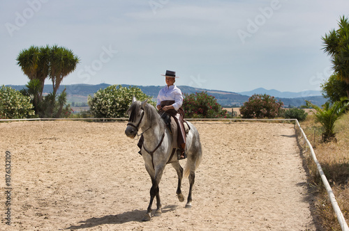 A young, beautiful Spanish woman dressed in riding clothes and wearing a traditional Spanish hat, riding a horse in the countryside on a sunny day.
