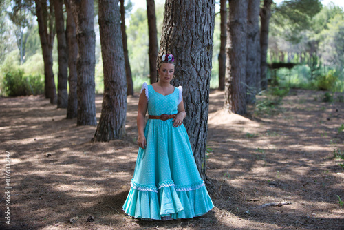 A beautiful young Spanish woman dressed in a traditional green flamenco dress and with a flower in her hair walks through the pine forests of Andalusia in the pilgrimage.