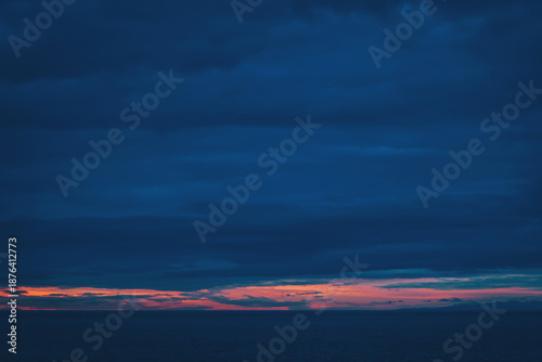 The sun sets over the ocean, casting orange and pink hues on the horizon. Dark clouds fill the sky, creating a striking contrast. The scene captures the end of the day by the water