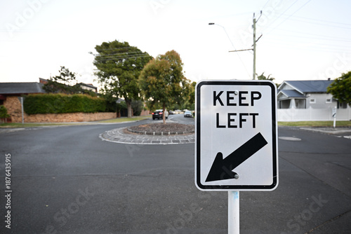 Rectangular white and black keep left traffic sign featuring a large arrow on a quiet suburban street, with roundabout and homes in the background