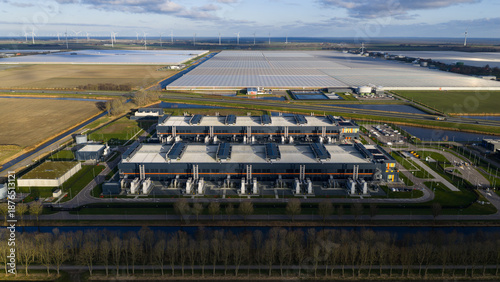 Aerial view of a vast data center contrasting with the shimmering greenhouses and distant wind turbines under the serene sky, Middenmeer, Netherlands.