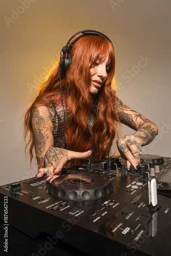 A female DJ is performing at a session. She has long red hair and tattoos. She is focusing on her equipment with headphones on. The setting appears to be well lit for a night event.