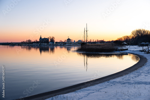 Winter sunset at Goitzsche Lake beach, snow on the shore, looking toward the promenade and marina, gauge tower close by, orange-red sky, capturing winter lakeside atmosphere in Central Germany.