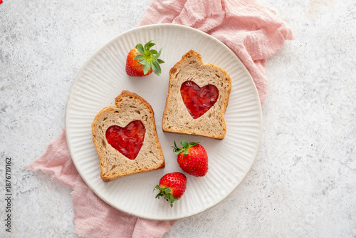 Toast with heart-shaped cutouts filled with jam and fresh strawberries on a plate with red heart decorations