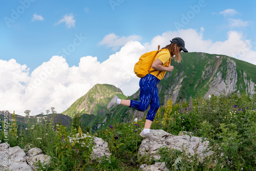 young woman traveler in cap with yellow backpack jumps from stone to stone on difficult rocky terrain during mountain hike in summer, hiking, adventure and exploration, active healthy lifestyle