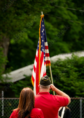 Back view of veterans in red shirts saluting American flag during patriotic ceremony or parade with green natural background. Shows military veterans honoring flag during Memorial Day, July 4, etc.