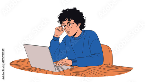 Serious young man with curly hair and glasses sits at a wooden table and works intently on a laptop computer in a clean and simple setting.