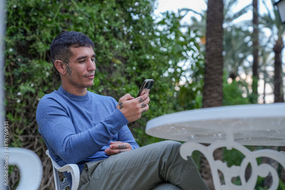 Obraz premium Smiling man with tattoos texting on phone while sitting in a garden