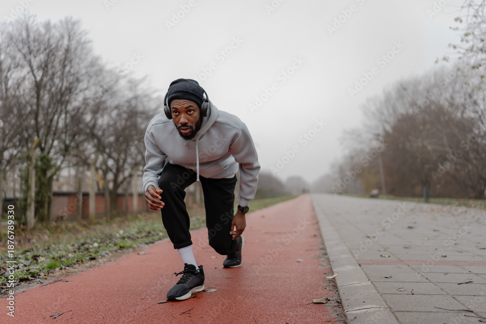 Fototapeta premium Focused athletic man in starting position on an outdoor running track, preparing to sprint. Urban fitness training, speed and power workout, healthy active lifestyle